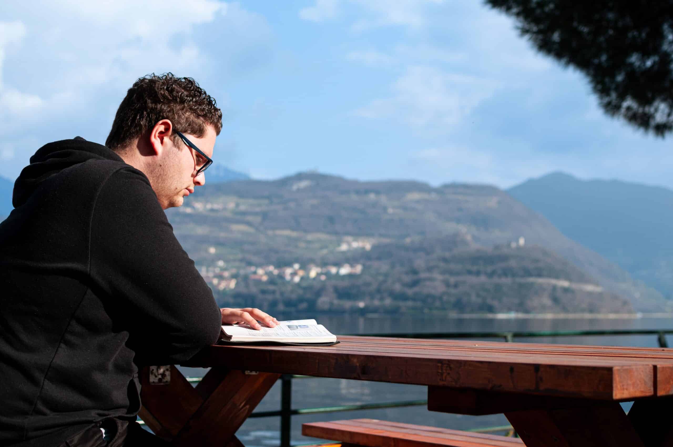 A man reading a book outdoors with scenic lake and mountain view in Milano, Italy.