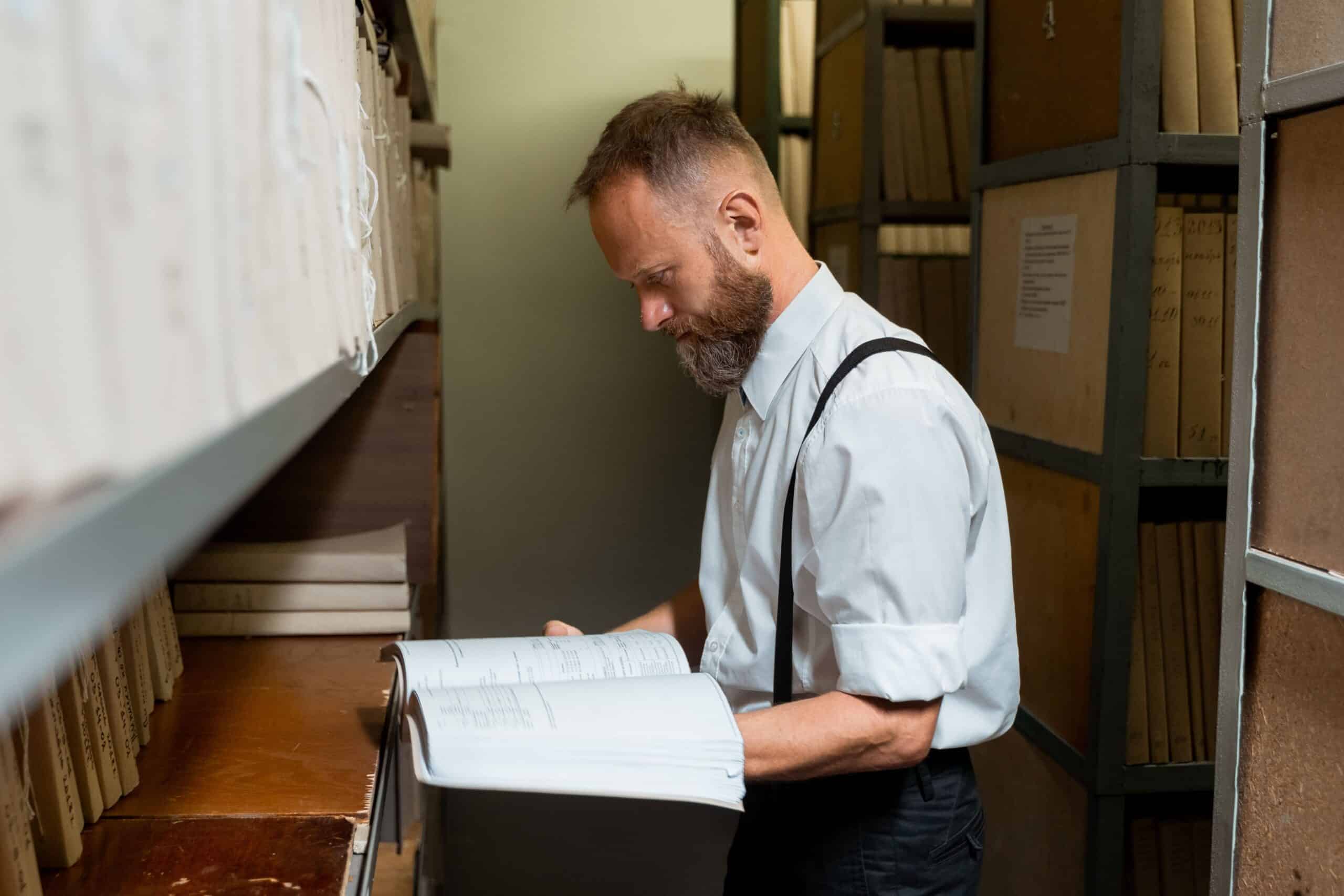 Bearded man in suspenders examining large book in archive room with wooden shelves.